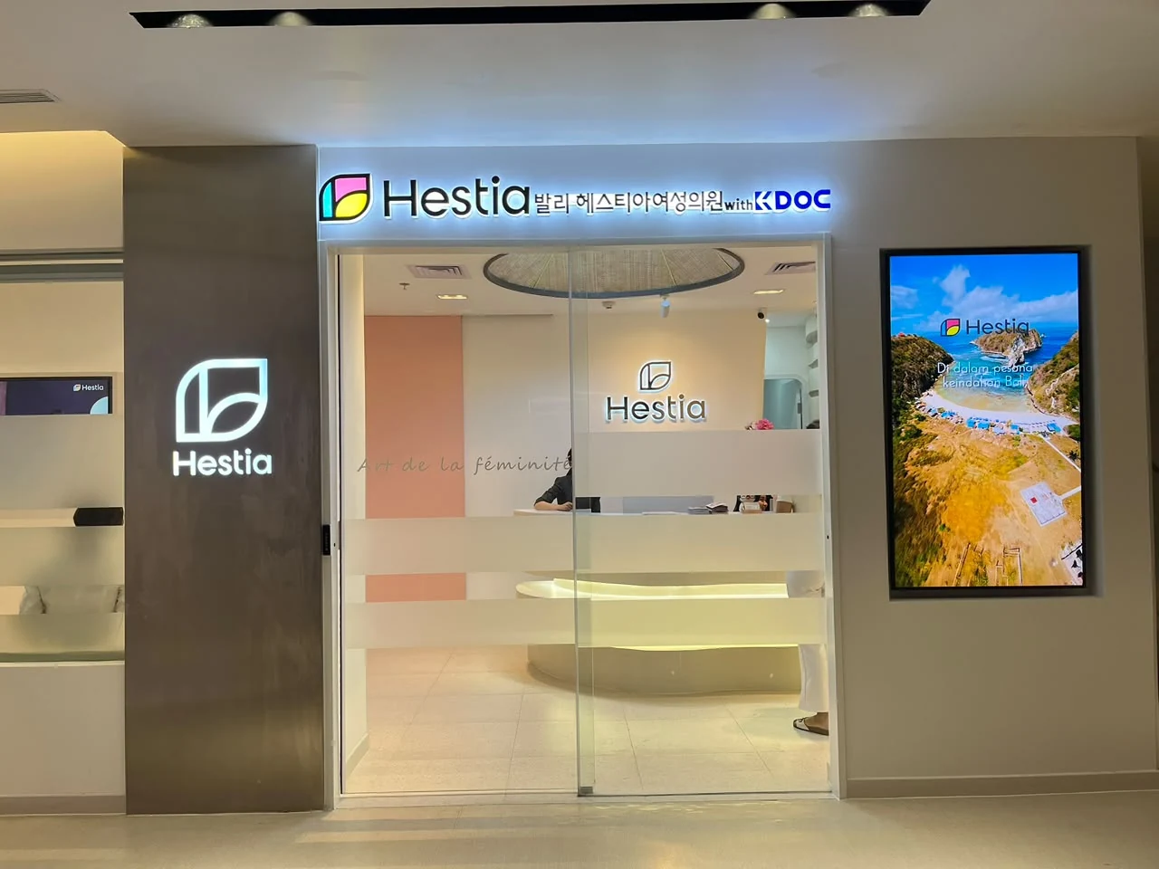 Entrance Hestia Women Clinic dengan pintu kaca, illuminated signage Hestia, digital display, dan area reception yang terlihat dari pintu masuk.