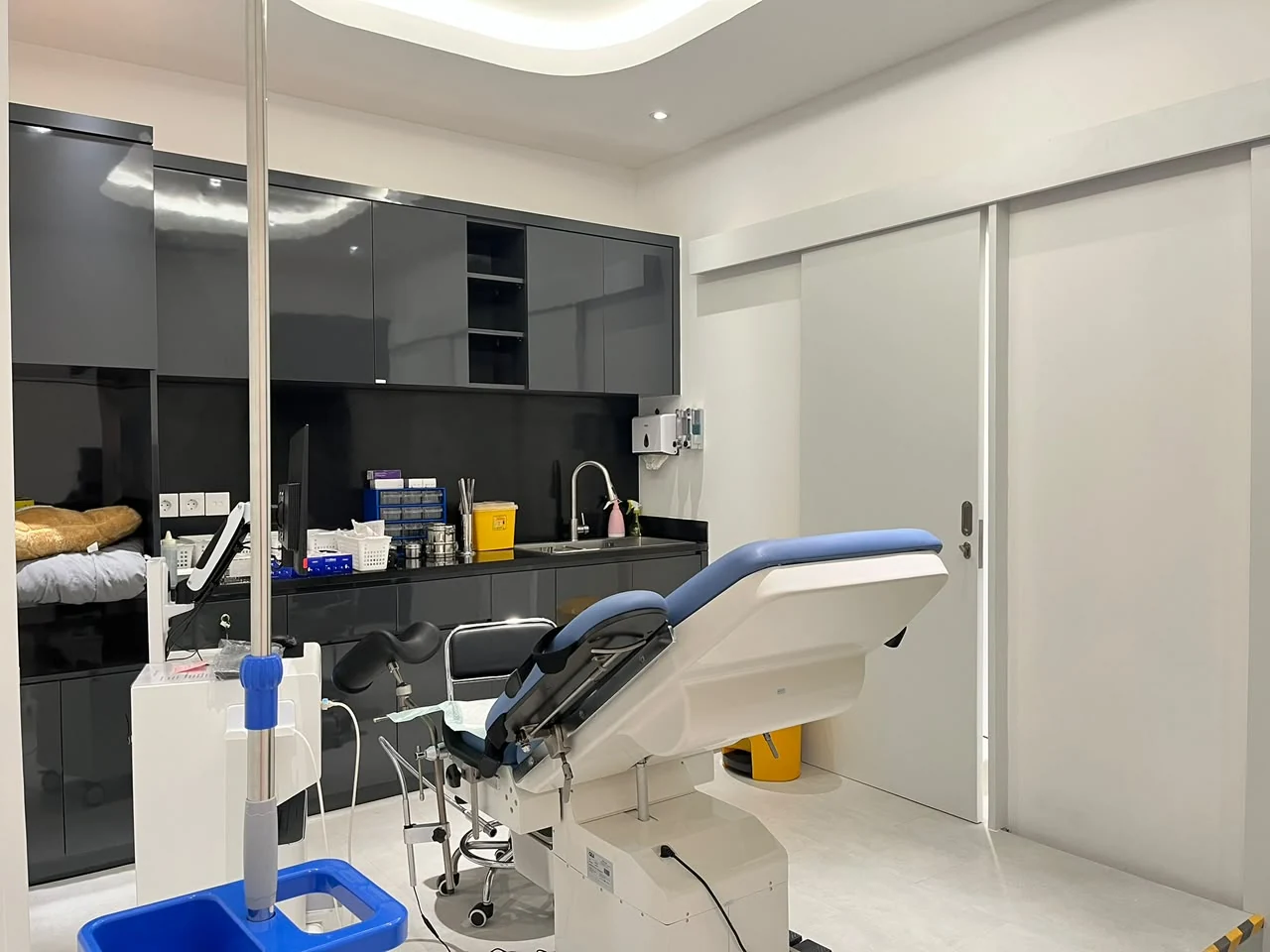 Ruang pemeriksaan Hestia Women Clinic dari sisi examination chair dengan custom cabinetry berwarna gelap, worktop, sink, peralatan medis, dan pintu geser.