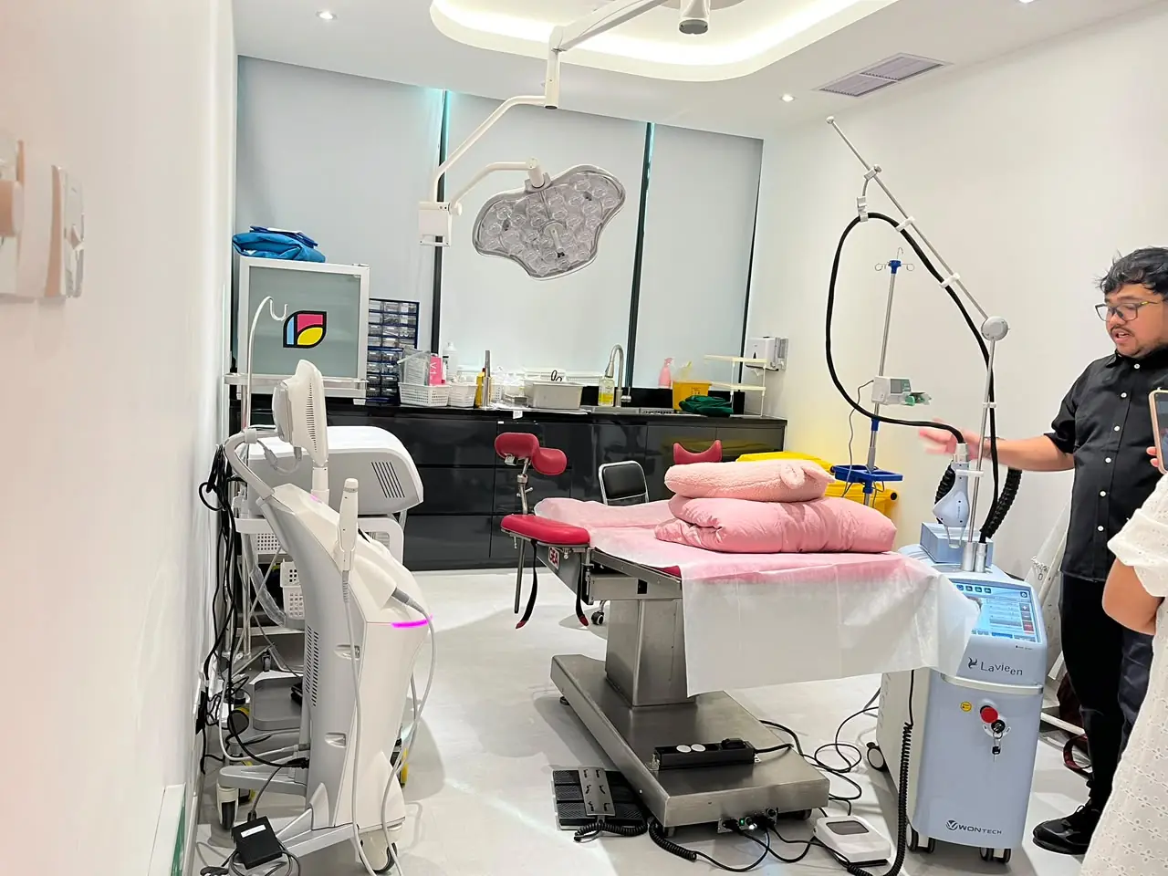 Ruang treatment Hestia Women Clinic dengan treatment bed, peralatan medis, custom cabinetry berwarna gelap, workstation klinis, dan overhead examination light.